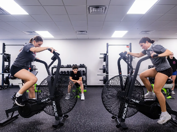 Two athletes conditioning on air bikes in the training floor